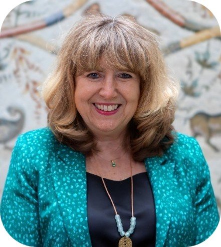 Portrait of Astrid Schomaker, Executive Secretary, Convention on Biological Diversity, smiling at the camera, with shoulder-length light brown hair and bangs. She wears a turquoise patterned jacket over a black top and two necklaces, one with pale blue beads and a gold-coloured pendant. A softly blurred decorative wall is behind her.