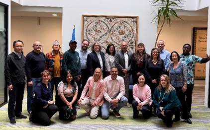 The Ad Hoc Technical Expert Group (AHTEG)  portrait of 19 people posing indoors in two rows, with 13 standing behind six kneeling or crouching in front. Most smile toward the camera. Behind them are a framed decorative textile, a partially visible blue flag, and a tall potted plant.